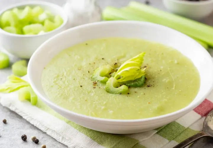 Zuppa cremosa di sedano verde, guarnita con gambi freschi e pepe nero. Accanto, una ciotola di sedano a cubetti.