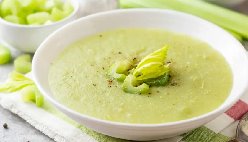 Zuppa cremosa di sedano verde, guarnita con gambi freschi e pepe nero. Accanto, una ciotola di sedano a cubetti.