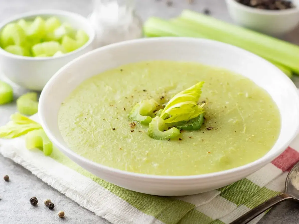 Zuppa cremosa di sedano verde, guarnita con gambi freschi e pepe nero. Accanto, una ciotola di sedano a cubetti.