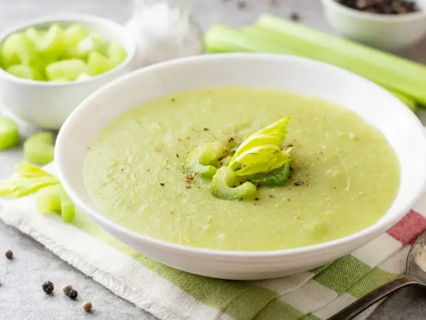 Zuppa cremosa di sedano verde, guarnita con gambi freschi e pepe nero. Accanto, una ciotola di sedano a cubetti.